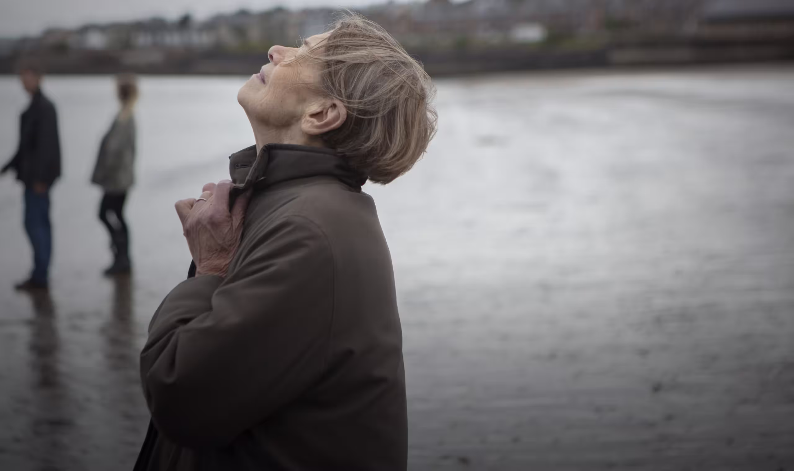 Elderly woman on a beach holding the collar of her jacket and looking in pain.
